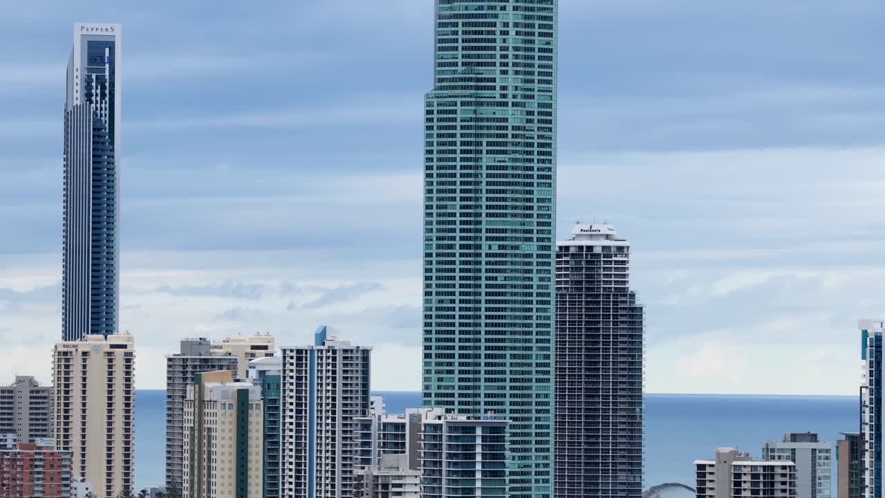 Drone footage captures the Gold Coast skyline, highlighting tall skyscrapers against a cloudy sky, with the ocean in the background