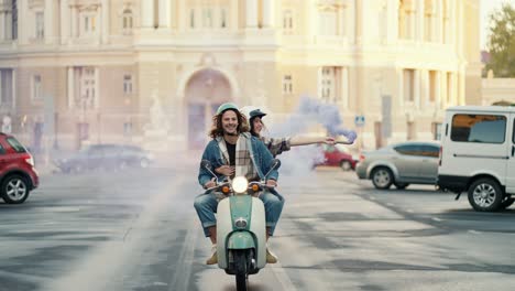 a happy guy with curly long hair in a denim shirt and pants rides a green moped with his girlfriend who is holding a blue fire in her hands that produces smoke and leaves behind a blue trail on a wide street in the morning in the city