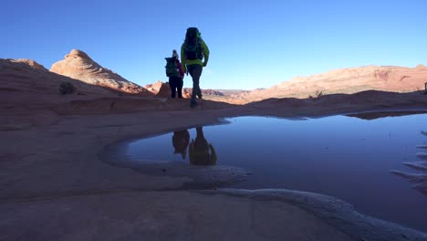 dos excursionistas con mochilas pasan junto a un charco de agua en un desierto rocoso