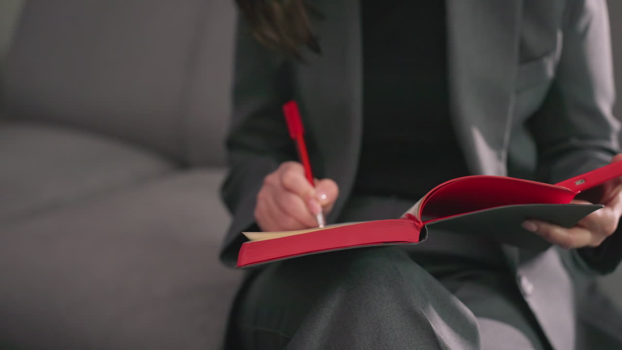 Businesswoman writing in red notebook with pen in hand, sitting in modern office, gray suit, focused on planning, decision-making, productivity, workspace, corporate environment