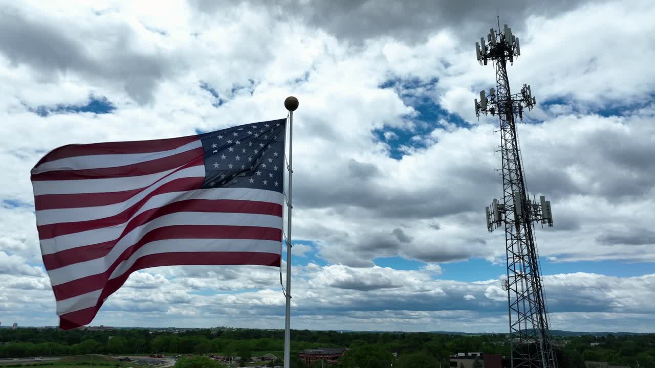 Proud American flag waving in wind. Aerial orbit shot. Spirit of USA. Television tower on cloudy day in American suburb. Modern connectivity in America.