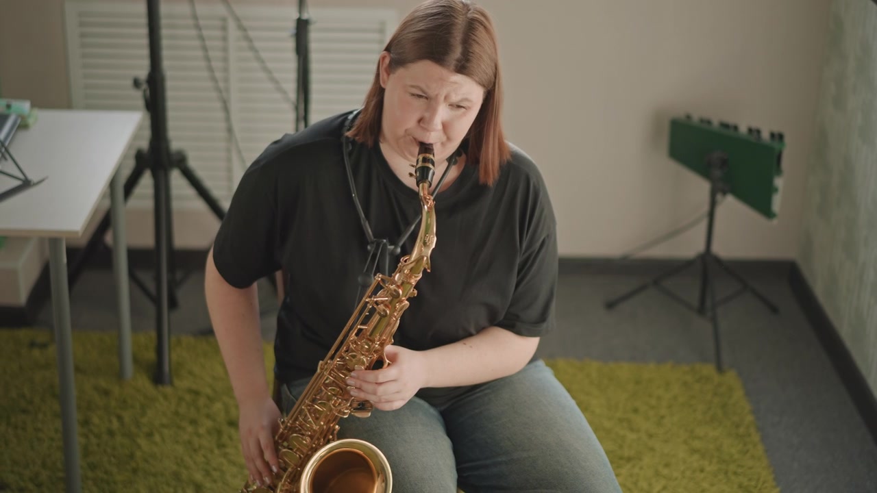 En artist övar känslosam saxofonimprovisation i en tyst skolstudio, med en coach som observerar utanför bild. Gröna akustikpaneler och keyboard i bakgrunden, i en fokuserad och uttrycksfull stämning.