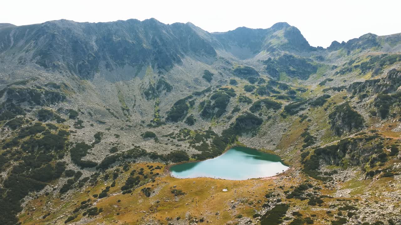 Blue glacial lake at the bottom of tall mountain ranges, surrounded by green pastures and mountain rocks, rising drone shot