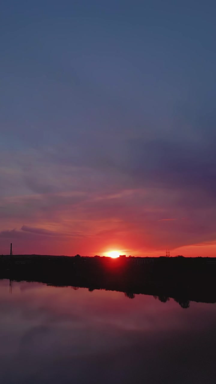 Vibrant orange sunset over river horizon with dark silhouette of city skyline. Purple and pink clouds reflecting in calm water during golden hour in picturesque landscape scenery.