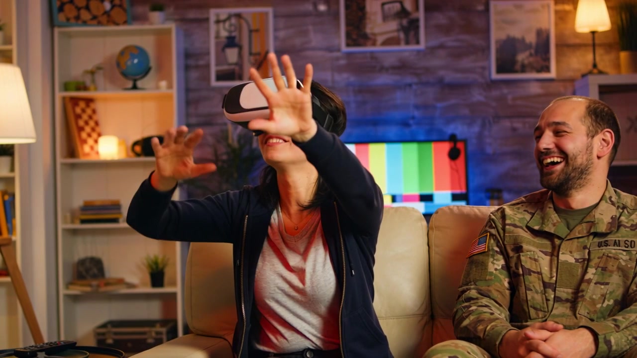 Donna in felpa con cappuccio che indossa un visore VR e si protende nella realtà virtuale a casa, mentre un uomo in mimetica la osserva.