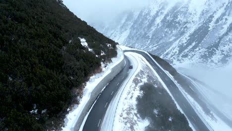 Vista-Aérea-De-Un-Coche-Pequeño-Conduciendo-Por-Una-Carretera-De-Montaña-Cubierta-De-Nieve-Durante-Un-Día-Nublado-De-Invierno-Con-Nevadas-En-Suiza