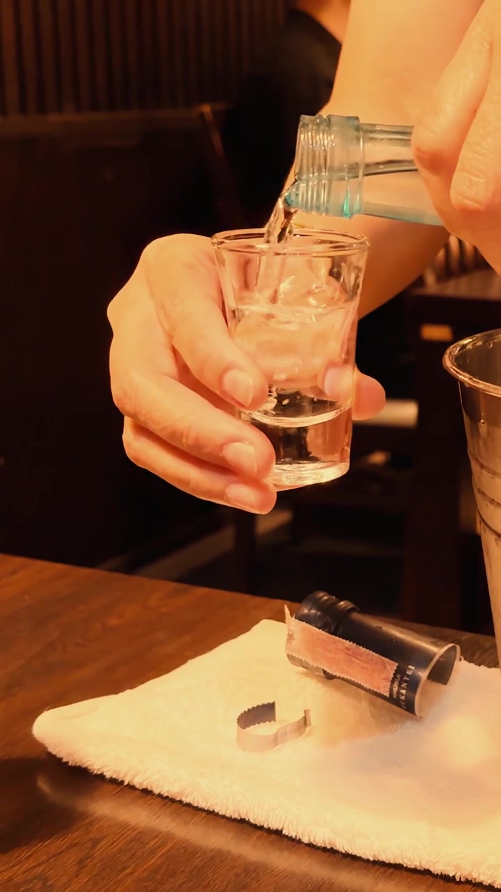 Primo piano di mani che versano alcol trasparente in un bicchierino da shot in un'illuminazione calda da ristorante.