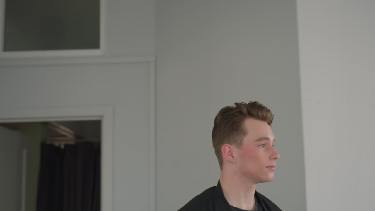 Young adult male with short hair walks out of apartment leaving door open stands near window holding travel bag looking thoughtfully into distance in softly lit minimal interior with neutral tones