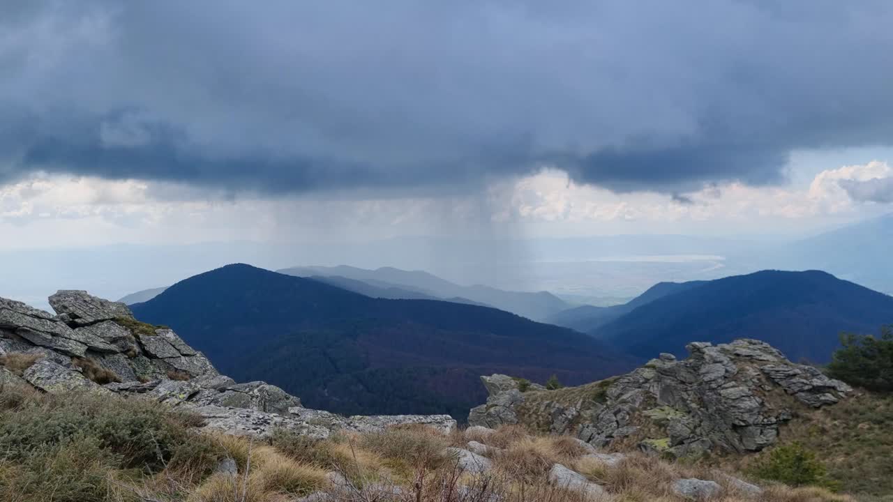 Udsigt til en bjergtop nær Lailias, Serres, Grækenland, under mørke stormskyer. Regn falder synligt på en fjern bjergryg, mens tunge skygger fejer hen over landskabet og fanger naturens rå, stemningsfulde skønhed.