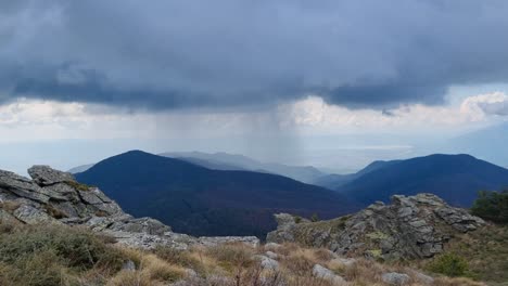 view of a mountain peak near lailias, serres, greece, under dark storm clouds. rain falls visibly on a distant ridge as heavy shadows sweep the landscape, capturing nature’s raw, moody beauty