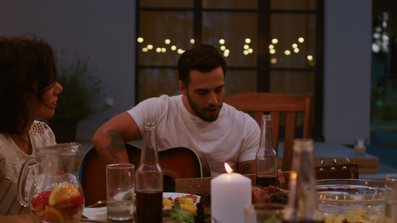 sentado en la mesa de la cena joven guapo toca la guitarra en la celebración de la fiesta del jardín de la noche de verano. amigos escuchando música conmovedora en un ambiente romántico.
