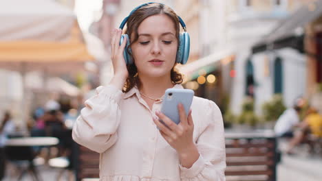 Happy-young-woman-in-wireless-headphones-choosing,-listening-music-dancing-outdoors-city-street