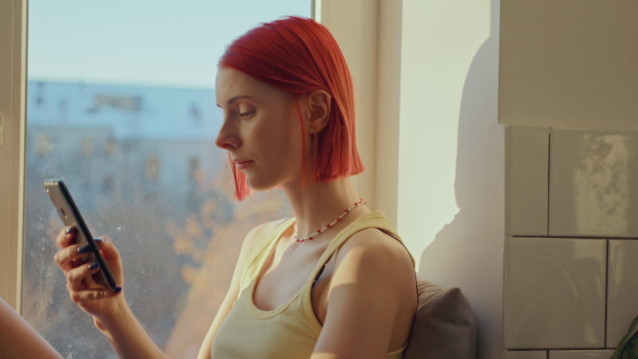 빨간 머리 소녀는 창문 앞에서 차가워서 밖을 바라보고 전화를 사용합니다.