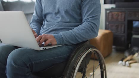 midsection of caucasian disabled man in wheelchair using laptop in living room