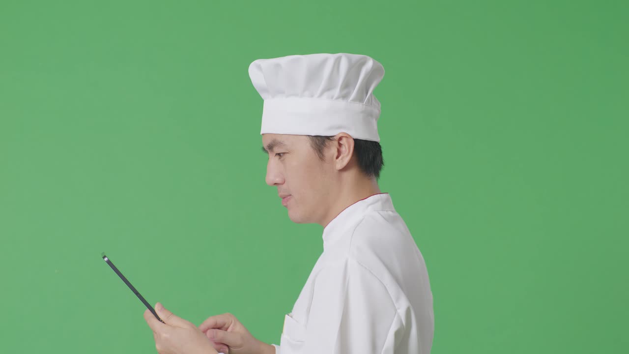 Vista laterale di uno chef asiatico che usa un tablet mentre cammina nello studio con sfondo green screen