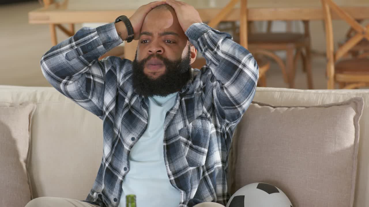 Vertical video: Reacting to off cue, man at home in plaid holding head on sofa with ball, bottle