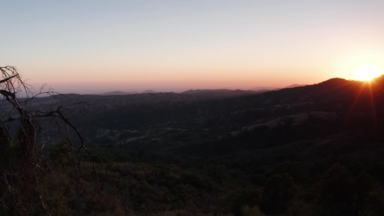 toma aérea volando más allá de un árbol en primer plano con el sol asomándose sobre una cresta de montaña al atardecer en el fondo