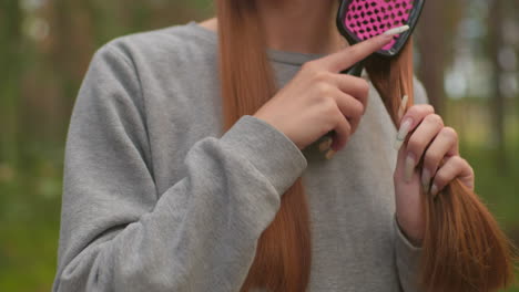 close-up of woman gently brushing her long, straight reddish-brown hair with a pink hairbrush, focusing on the smooth texture and care in a serene forest setting