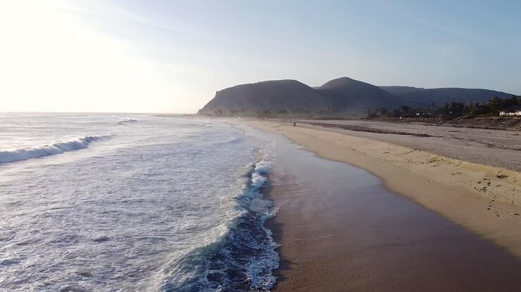Close up of waves and ocean mist crashing into sandy beach during golden hour sunset with drone flyover