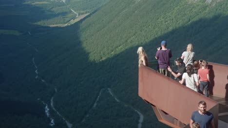 chica rubia de pie en una plataforma con un montón de turistas en el sol y disfrutando de la vista del trollstigen en noruega