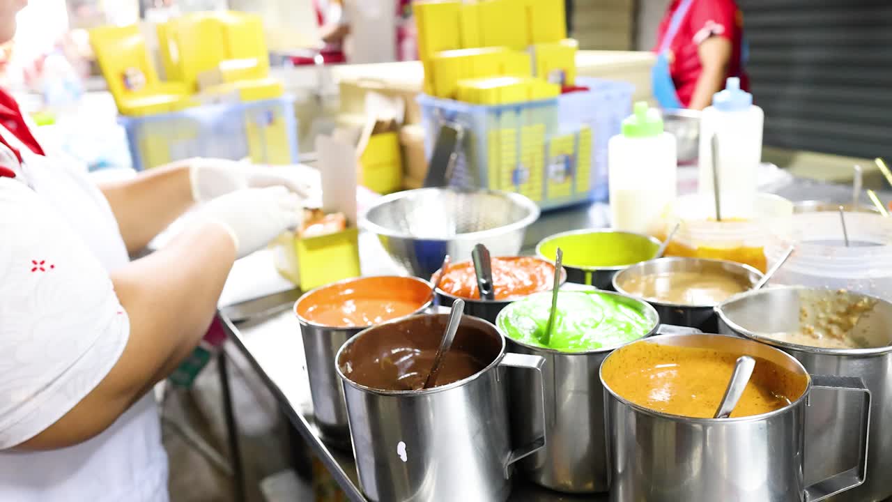 Vendor wearing gloves spreads colorful sweet pastes on bread at vibrant Bangkok street food stall