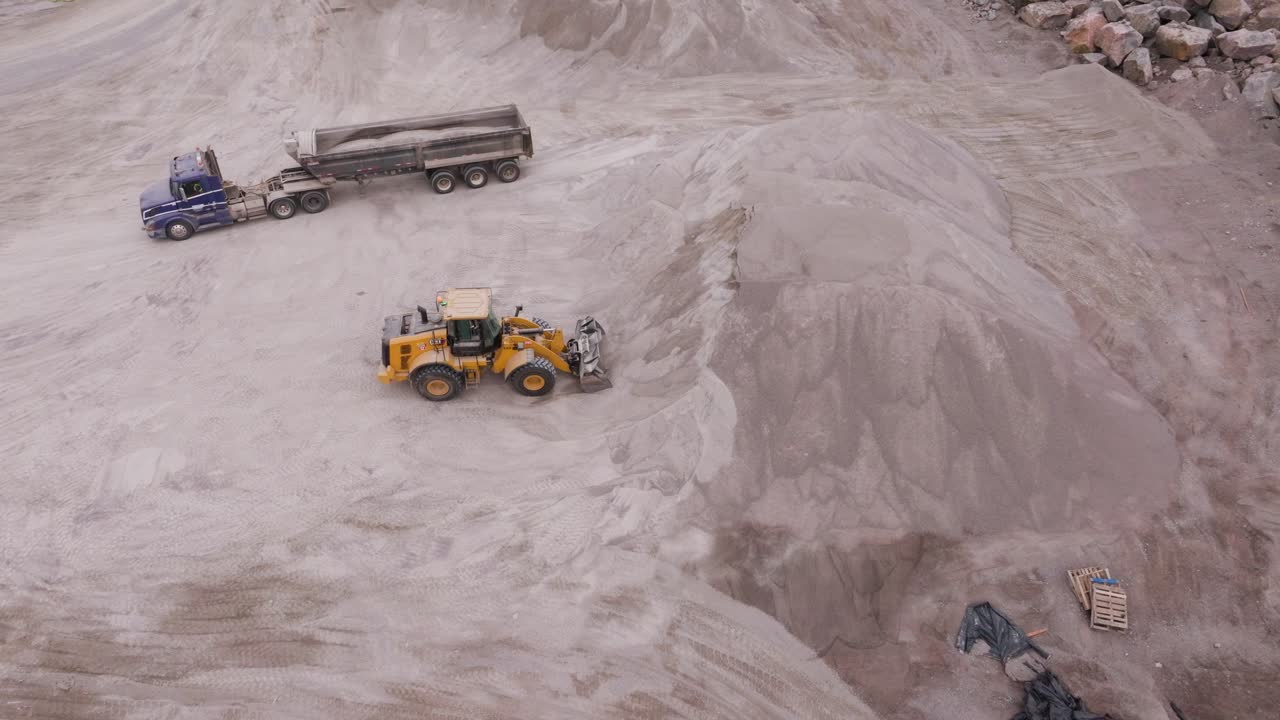 Aerial drone footage of an open-pit mining operation surrounded by dense forest on a sunny day. Heavy machinery and haul trucks working in a large industrial site