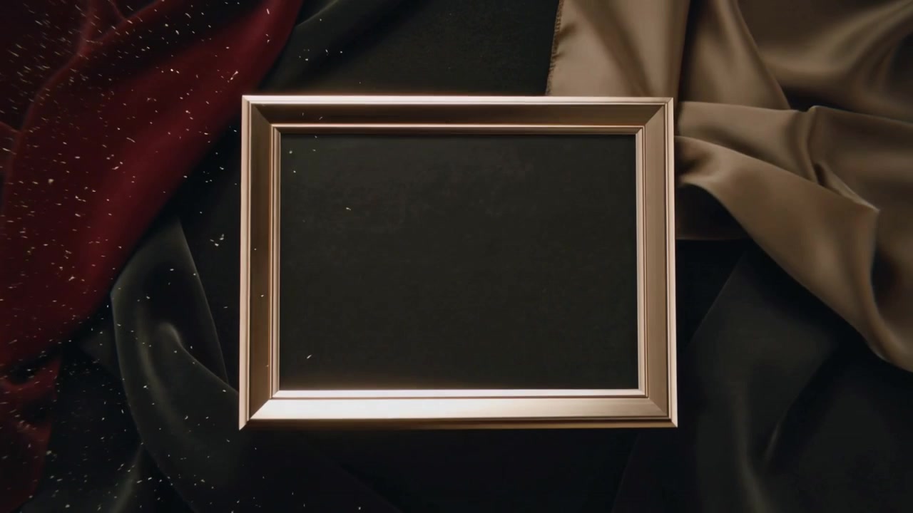 Rotující kovový rám začíná nakloněný, pohybuje se po saténovém stole, třpytky se rozptylují blízko levého okraje.