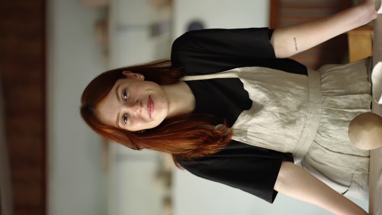 la alfarera de cabello rojo sonríe, sus productos están cerca de ella contra el fondo del lugar de trabajo