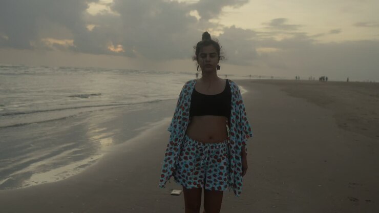 An Indian woman gracefully walks along the beach, nearing the camera with the sea as her tranquil backdrop