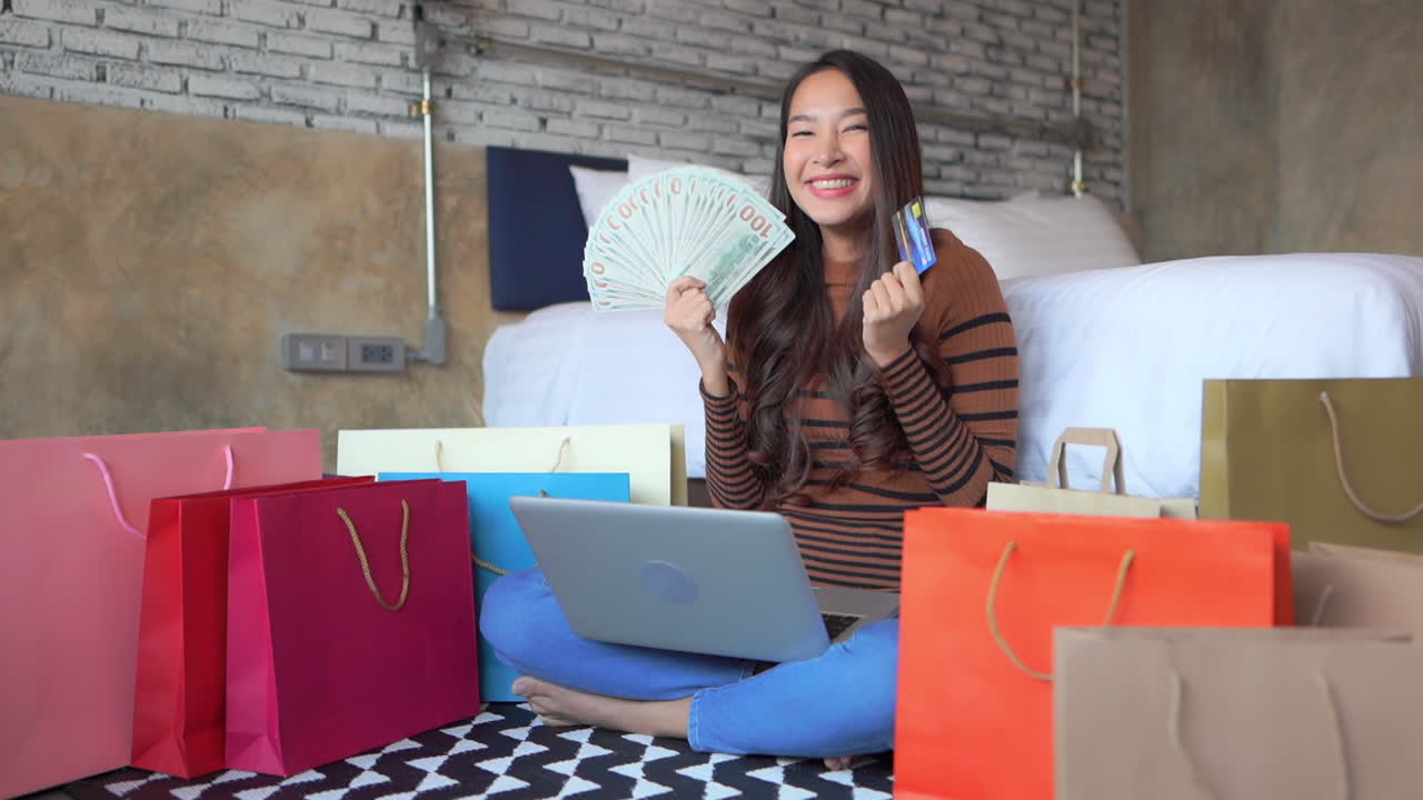 Pretty Asian woman holding a big pile of money in her hands, she won the prize, lottery, online casino, shopping money, sitting on the floor with a notebook