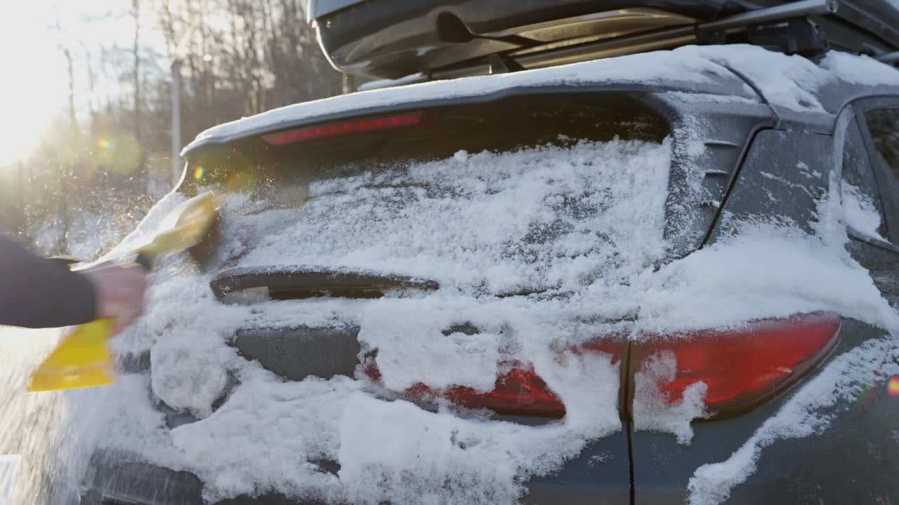 Man uses brush, scraper to clear snow and ice from rear window of SUV on winter morning in Norway.