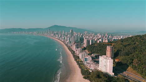 drone image showing the sweeping shoreline, tall buildings, and forested hills of itapema and meia praia, in the state of santa catarina, brazil, with calm ocean and clear sky in the background