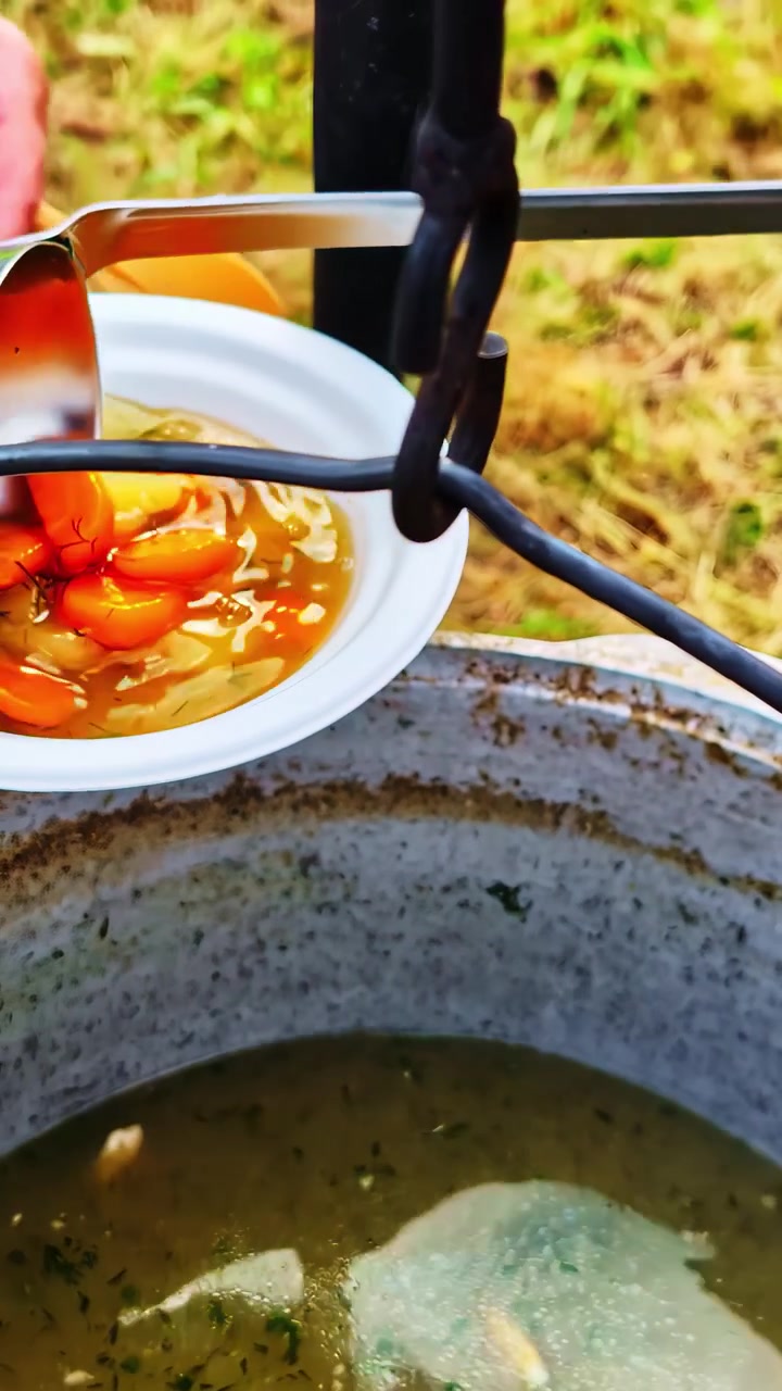 Un mestolo versa una zuppa sostanziosa in una ciotola da una grande pentola all'aperto, creando un'ambientazione rustica per il pasto.