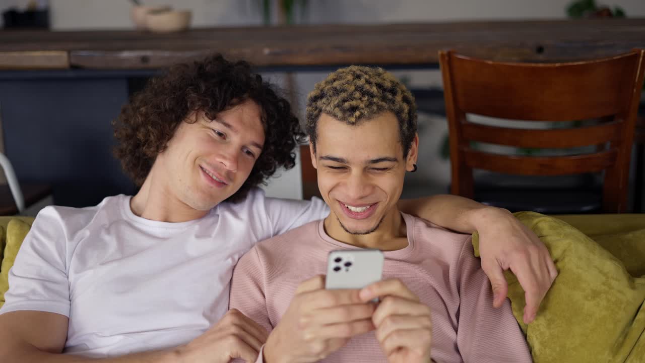 pareja gay relajándose en el sofá y abrazándose, viendo la pantalla del teléfono inteligente