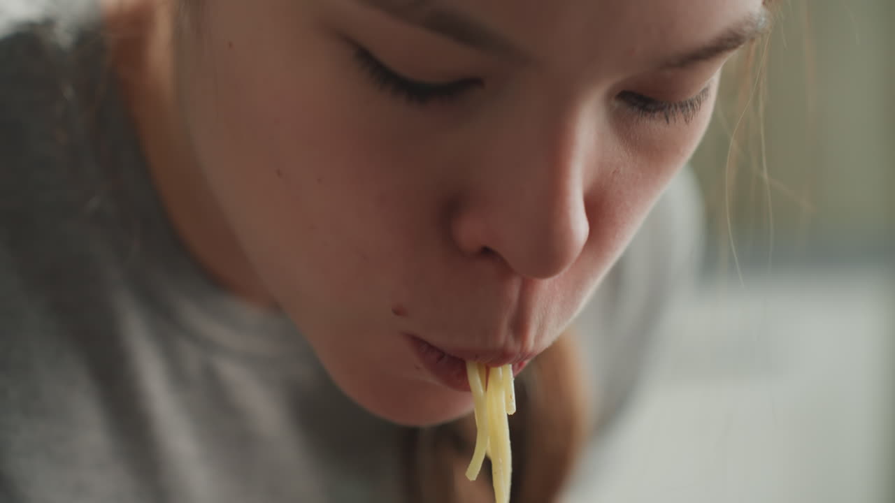 Seorang wanita makan mi. Mahasiswa menikmati makanan cepat saji. Wanita di dapur dengan rambut dikepang makan mi dengan gembira. Mahasiswa fokus dengan rambut dikepang menikmati hidangan mi panjang yang menenangkan