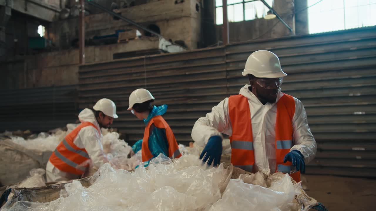 A man with Black skin in a white protective uniform in an orange vest together with his colleagues sorts and presses plastic cellophane garbage at a waste processing and sorting plant
