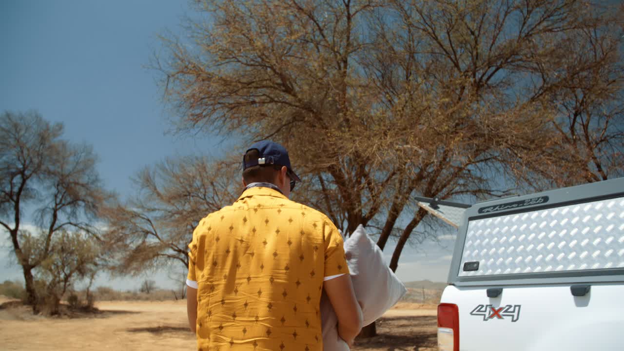 Hand held tracking shot of a off-roading Caucasian male tourist in Africa as he climbs out of his rooftop tent and then proceeds to carry his pillows out of his tent to the trunk of his vehicle.