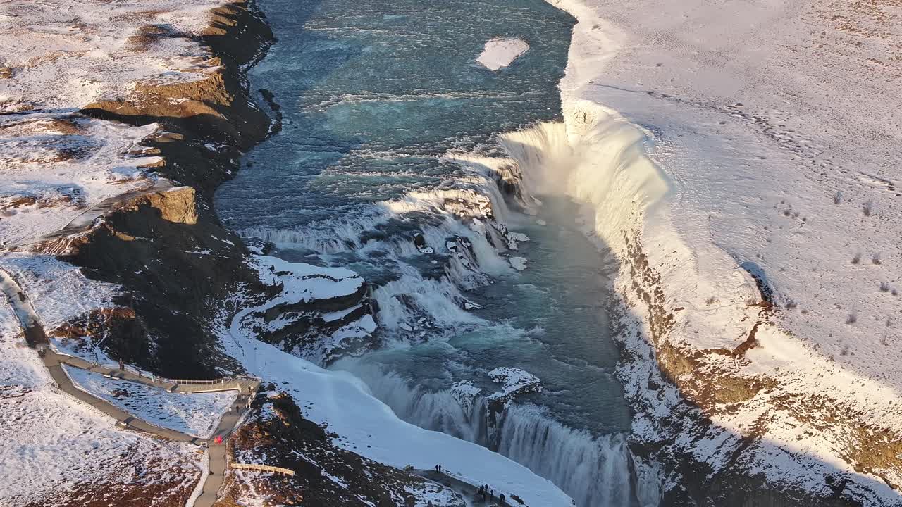 Majestic aerial view of Gullfoss waterfall in Suðurland, Iceland, where the Hvítá River plunges into a rugged canyon surrounded by snow-covered cliffs.