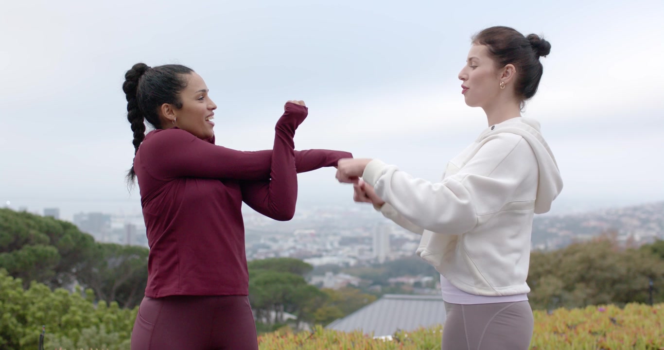 Diverse female duo in gear on hillside above city, instructor reaching, guiding arms for stretch