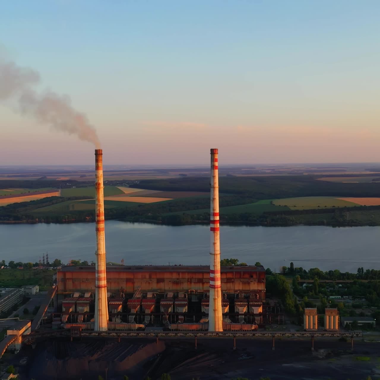 Manufacture near the river. Pipes with dirty smoke on nature background in the evening. Industry among nature at sunset. Aerial view.