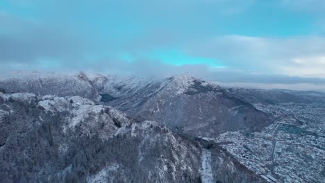 Bergen-Panoramic-aerial-drone-ascends-above-port-town-and-harbor,-Norway-covered-in-snow