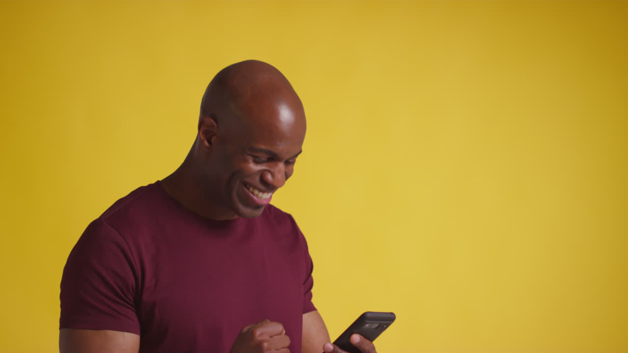 Studio Shot Of Excited Mature Man With Mobile Phone Celebrating Winning Money Against Yellow Background