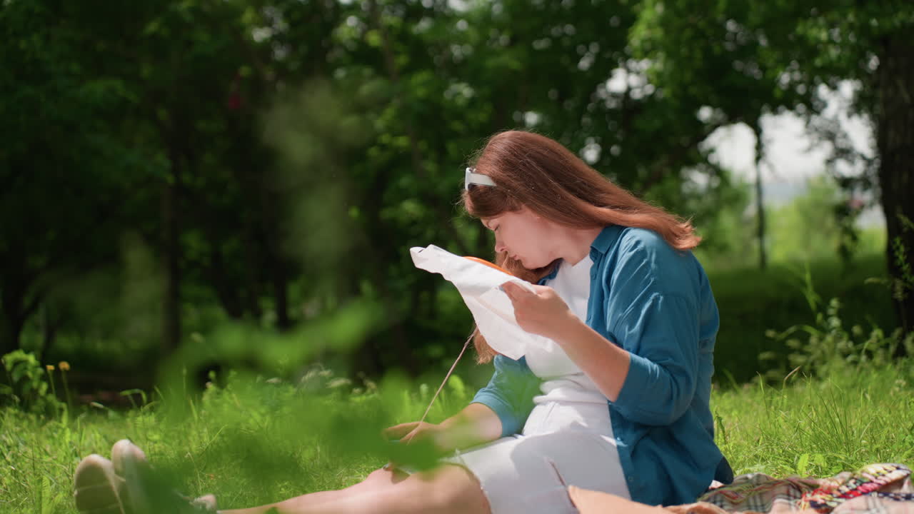 Close up of lush greenery with lady watching something attentively then continuing embroidery on white fabric under sunlight in calm park, expressing focus, patience, and creative dedication