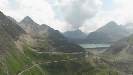 aerial view of winding mountain road grimsel pass with wind turbines on ridge line in background beside dam