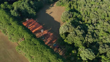 Aerial:-outdoor-red-clay-tennis-courts,-tree-shadows-over-sport-complex