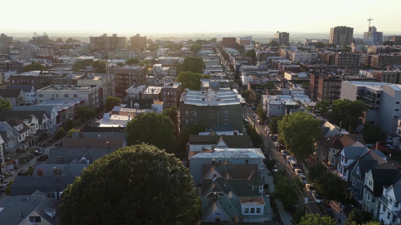 Case vittoriane con tetto a capanna e villette a schiera in mattoni costeggiano una tranquilla strada residenziale verso grattacieli distanti, alberi fitti risplendono con il tramonto dorato retroilluminato a Union City, New Jersey. Ripresa aerea con drone.