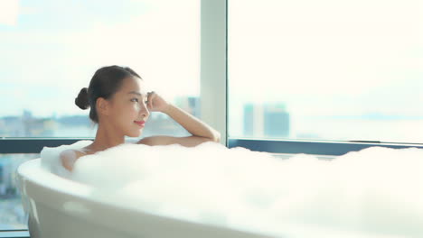 편안하게 목욕을 하는 아시아 여성, 그녀는 배경에 유리 벽이 있는 고급 아파트 단지의 높은 층에 거품이 가득한 욕조에 누워 있습니다.