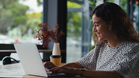 Joven-Empresaria-Sentada-Con-Una-Computadora-Portátil-En-Una-Cafetería-Tomando-Una-Llamada-Por-Teléfono-Móvil-Usando-Un-Micrófono