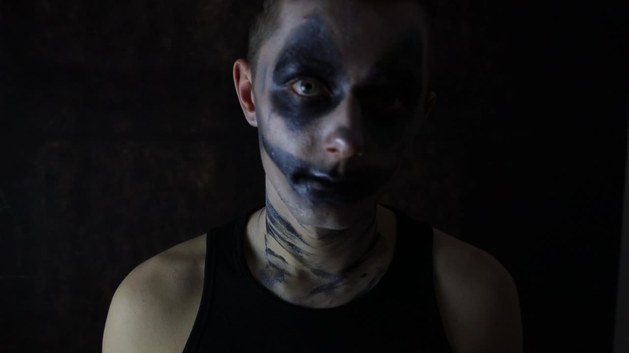 Young man with a scary Halloween dark clown makeup standing in front of a black background