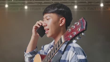 vista lateral de cerca de un adolescente asiático con una guitarra hablando en un teléfono inteligente mientras está de pie en el escenario